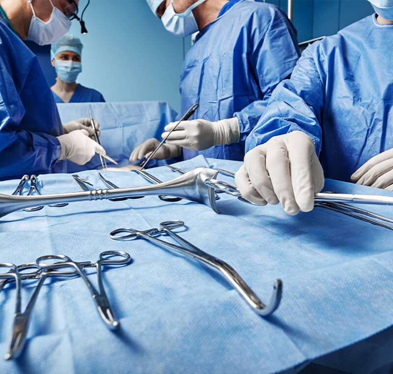 A surgical technologist in full sterile gown, gloves, and mask stands at an operating-room instrument table, selecting and passing surgical tools to the surgeon during a procedure.